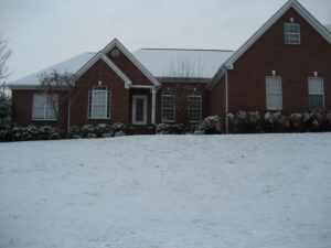 A house with snow on the ground and bushes in front of it.