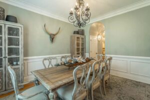 A dining room with a table and chairs, a chandelier and a wall mounted deer head.